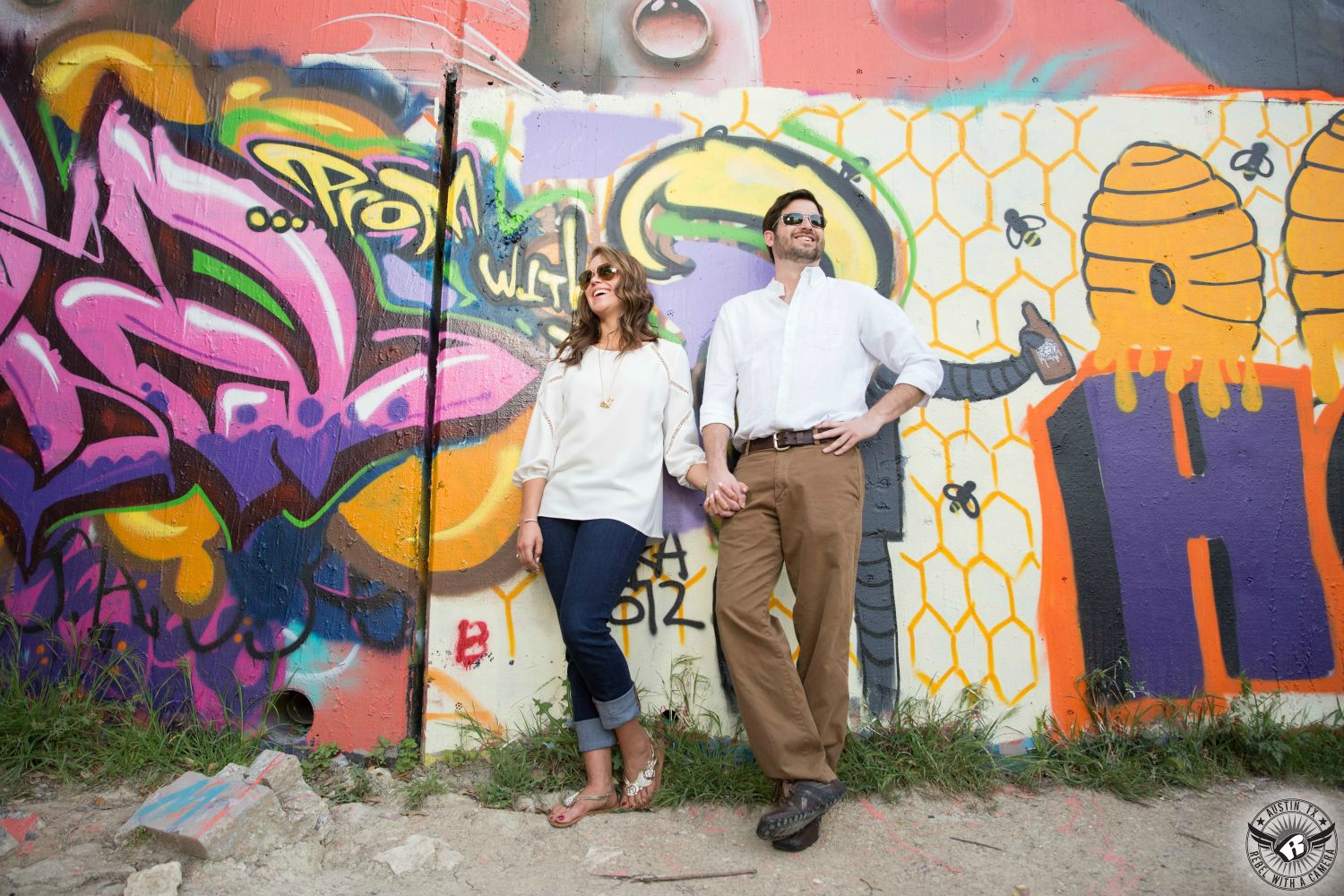 engagement couple with sun glasses laughing in white shirts and jeans with flip flops leaning against a wall covered in graffiti with bright colors and a honeycomb and bender and words all over the wall 